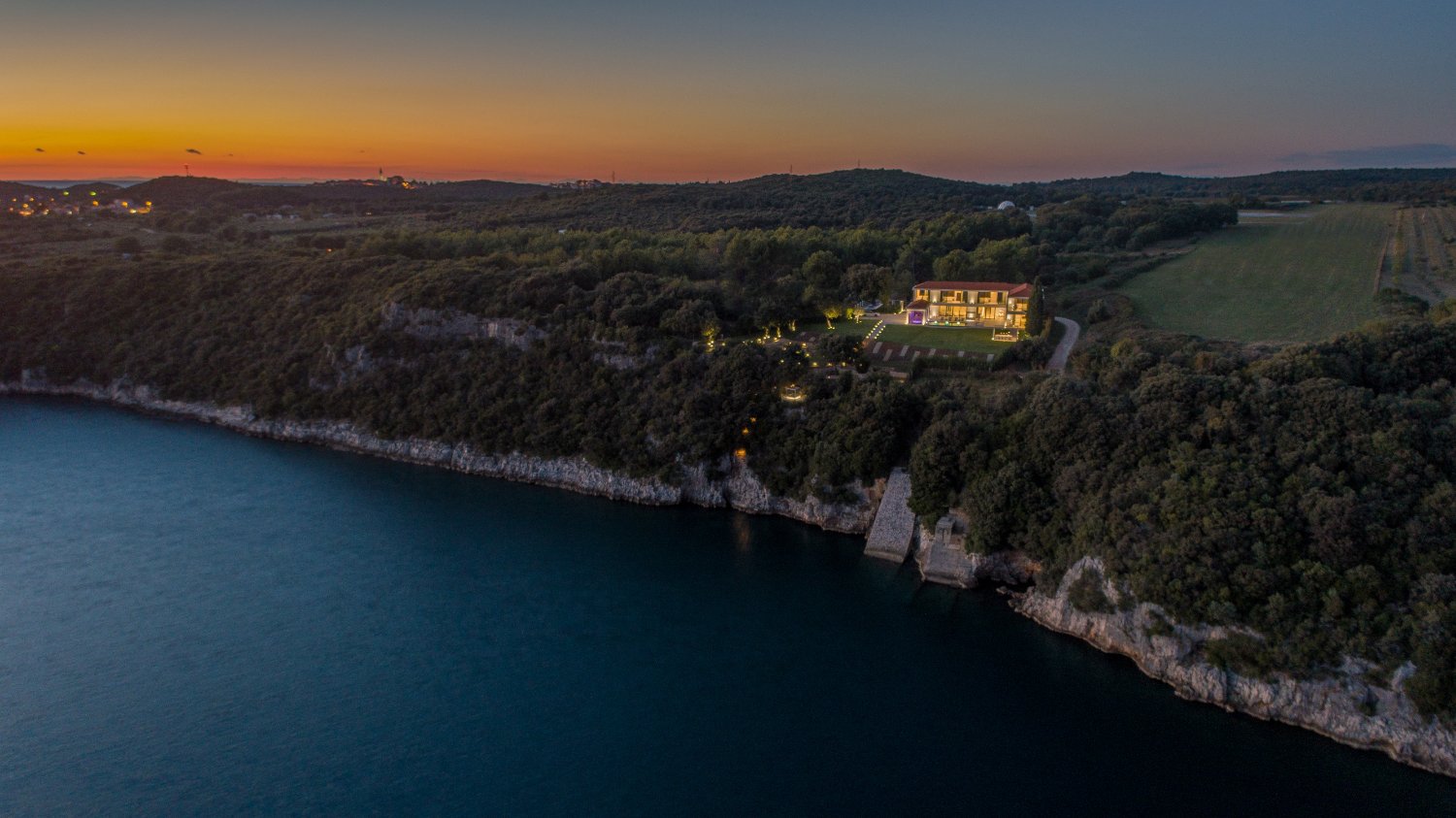 Zračna snimka luksuzne vile na litici uz more tijekom sumraka s osvijetljenim eksterijerom i pogledom na zalazak sunca.